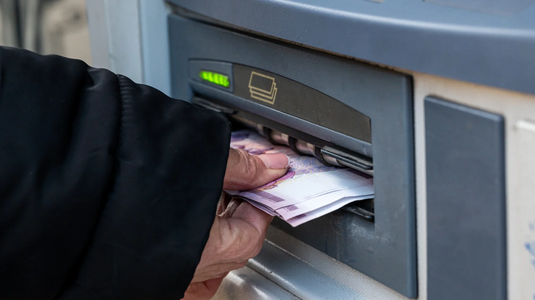 Bankautomata előtt készpénzt felvevő kéz, bankjegyekkel.